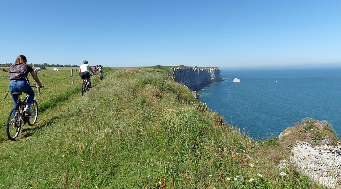 A trail of cyclists ride along a grassy path on top of the cliffs at Etretat